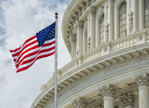 A government building with an American flag outside and in front of the building. A portion of the cloudy sky can be seen in the background.