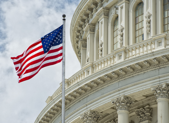 A government building with an American flag outside and in front of the building. A portion of the cloudy sky can be seen in the background - squared off.