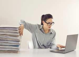 A woman is sitting a computer desk next to a large stack of documents. She has a lap top computer open on the desk and is looking at the computer screen while typing.