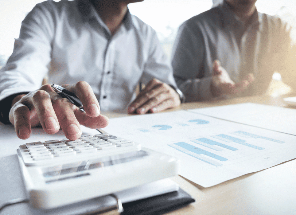 Two investors are sitting next to each other at a desk looking over documents. One of the people is types on a calculator while the other gestures toward the documents on the desk.