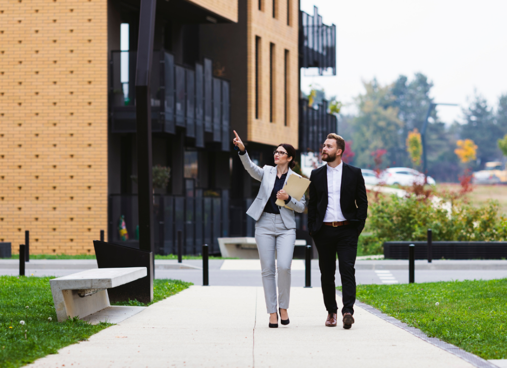 A real estate professional is showing an apartment complex to an investor. Both individuals are wearing business suits. They are walking through the apartment complex and evaluating the buildings.