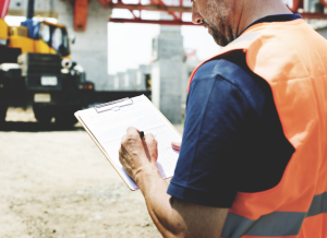 A construction worker is standing at a construction site and holding a clipboard and pen. He is looking down at the clipboard in his hand and writing on a document with his pen. The worker is wearing a hard hat and orange vest.
