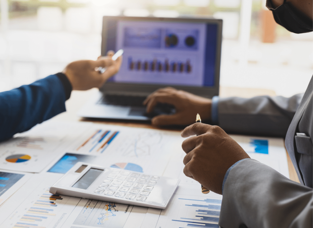 Two business persons are sitting at a table and pointing toward an open laptop computer. There are documents, graphs, and a calculator on the table. The laptop screen is also showing a graph.