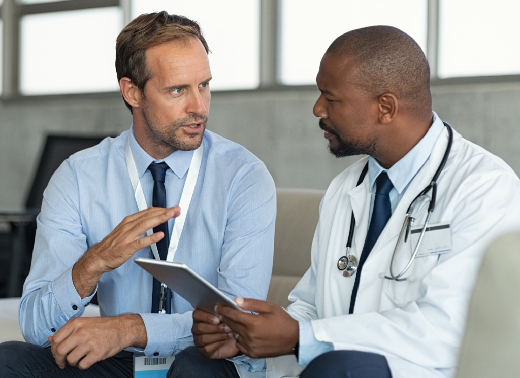 A medical doctor is speaking with another professional, who is wearing business attire and a name bade. He is looking at the doctor and talking, while the doctor listens intently. The medical doctor is wearing a white coat and stethoscope, and is holding a tablet computer.