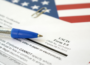 A blue pen is on top of a form I-9, which held together by a paper clip. The the blue pen and form I-9 are on top of a United States flag, which is partially shown in the blurred background.