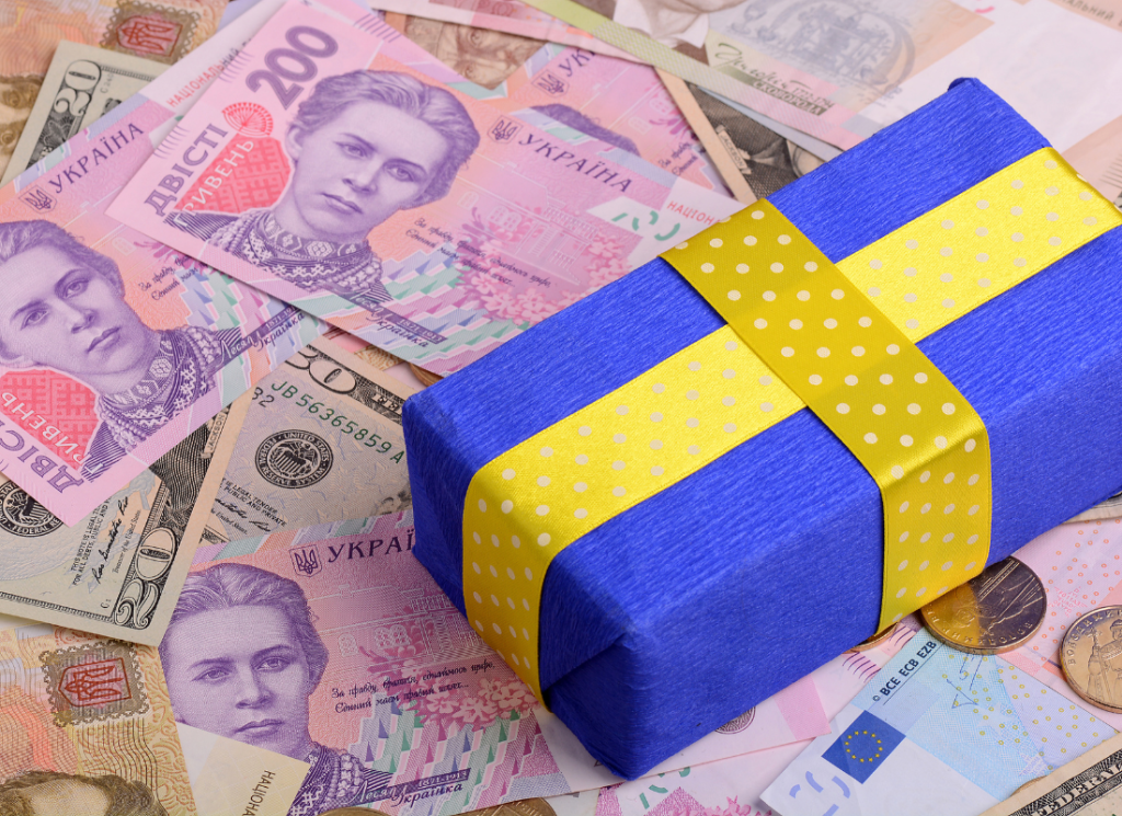 A gift wrapped in blue and yellow wrapping paper is sitting on top of money. The money is mostly U.S. and foreign cash, with a few foreign coins.