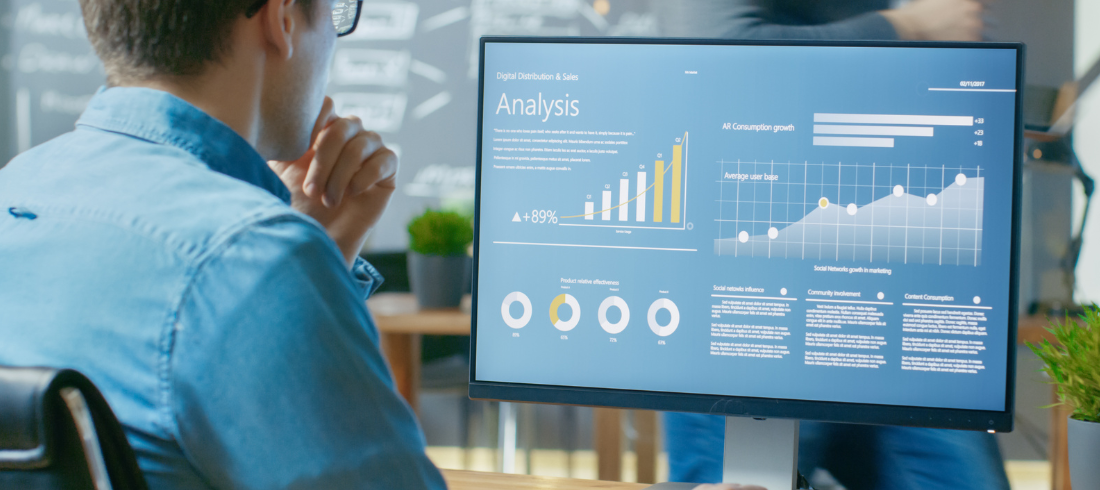 A worker in a office setting is viewing an analysis on a desktop computer. Multiple graphs are displayed on the computer screen. A second person can be seen in the background along with a blackboard with writing.