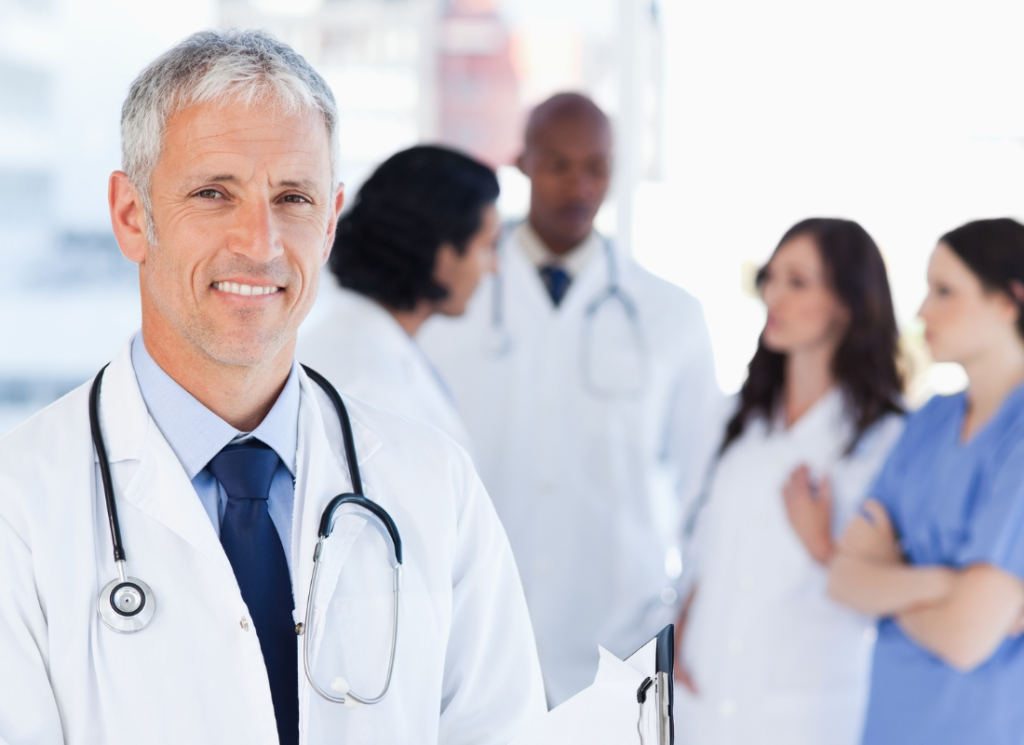 A medical doctor, wearing a white coat and stethoscope, is standing facing forward and smiling. In the background, four other healthcare professionals are having a discussion behind the doctor.