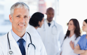 A medical doctor, wearing a white coat and stethoscope, is standing facing forward and smiling. In the background, four other healthcare professionals are having a discussion behind the doctor.