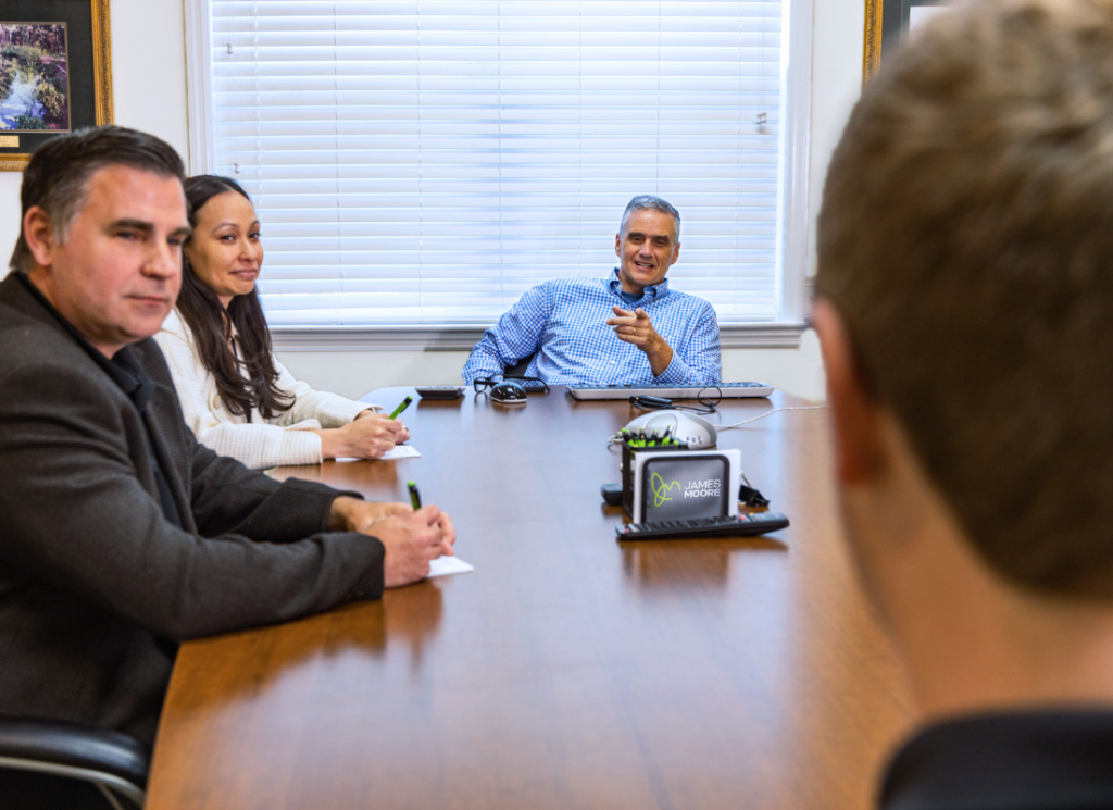 James Moore members are gathered at a table in a conference room. The leader of the group is pointing across the table at an employee and smiling. The other two team members at the table are also looking and smiling at the employee.