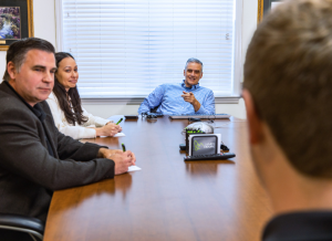James Moore members are gathered at a table in a conference room. The leader of the group is pointing across the table at an employee and smiling. The other two team members at the table are also looking and smiling at the employee.