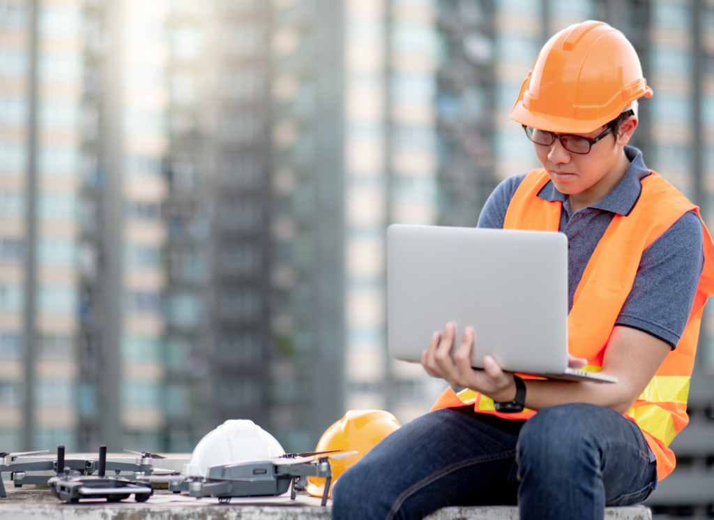 A construction worker is working on a laptop computer at a construction site. The worker is sitting down outside at a construction site and high rise buildings are in the background.