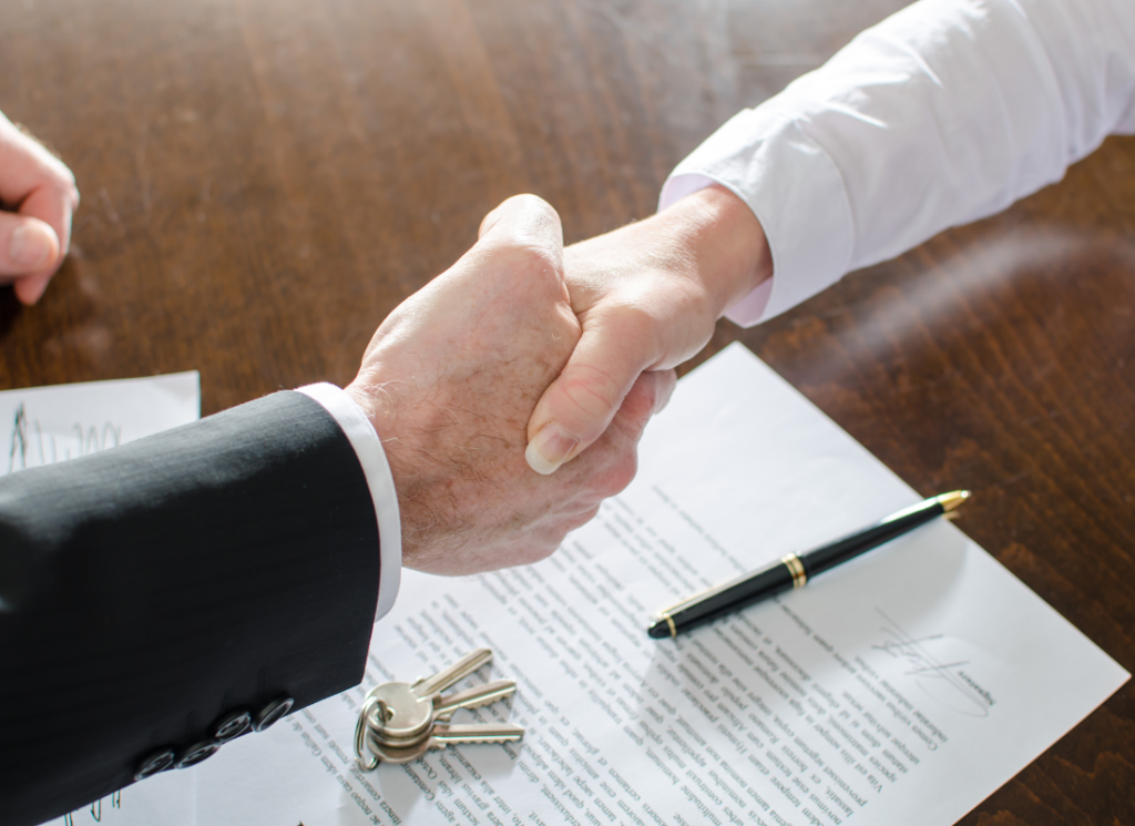 Two people are shaking hands accross a desk. There is a contract on desk with a pen and keys laying on top of the signed contract.