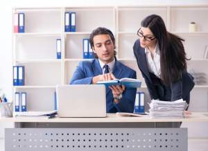 A business man is sitting at a computer desk, holding up an employee handbook, and pointing at a page in the book to show his co-worker. The co-worker is standing next to him and leaning over his shoulder to read from the handbook.