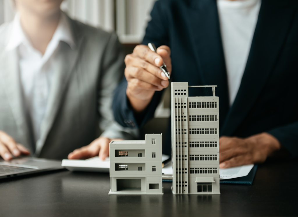 A real estate professional is sitting at a desk and examining a model of a building. A second person in the background is sitting at the same desk and typing on a calculator.