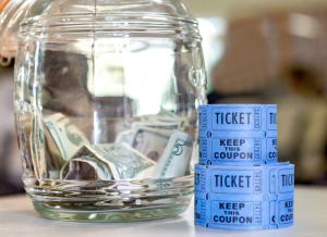 Two roll of raffle tickets are stacked together on a table next to a jar that is partially filled with cash.