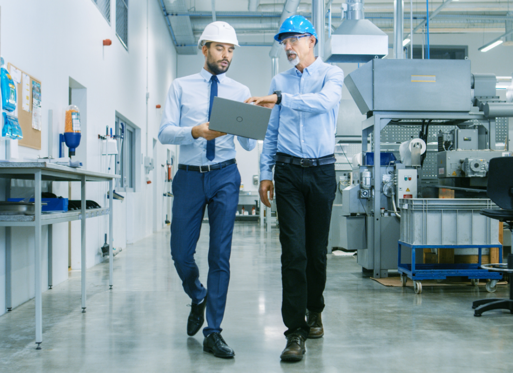 Two business men are walking in a factory and wearing hard hats. One of the men is holding a laptop computer and the other is pointing at the screen.