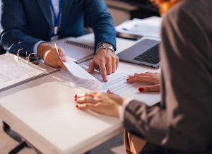 Two people wearing business attire are sitting across from each other at a desk and reviewing a contract. One individual is pointing to a specific line of the document or contract.