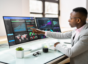 A businessman at a computer desk is pointing at a Data Analytics Dashboard displayed on his computer monitor.