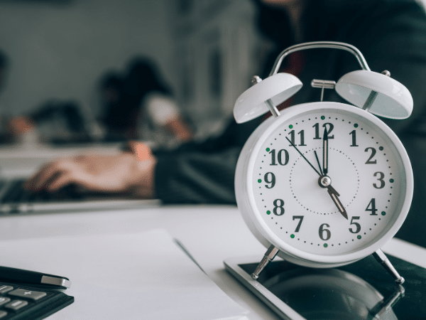 A clock showing 5 o'clock in the evening as employees are working in an office in the background.