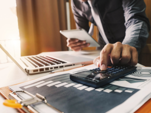 An accountant is standing at a desk while typing on a calculator and holding a notepad. There is an open laptop computer and clipboard on the desk.