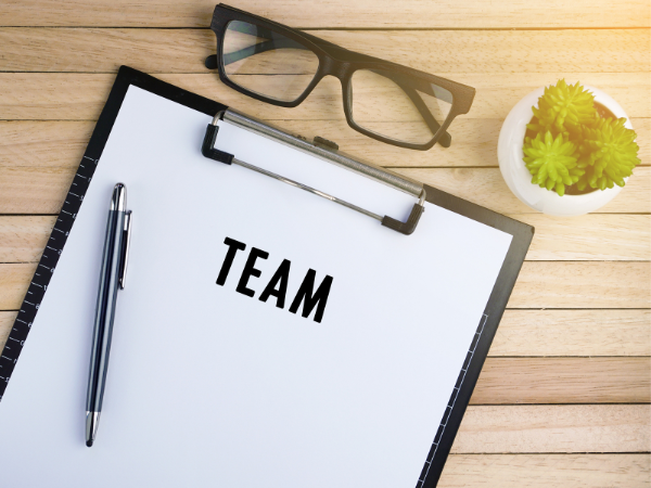 A blank list on a clipboard with the heading "TEAM". A pen sits on the clipboard. A pair of glasses and a small plant is near the clip board.