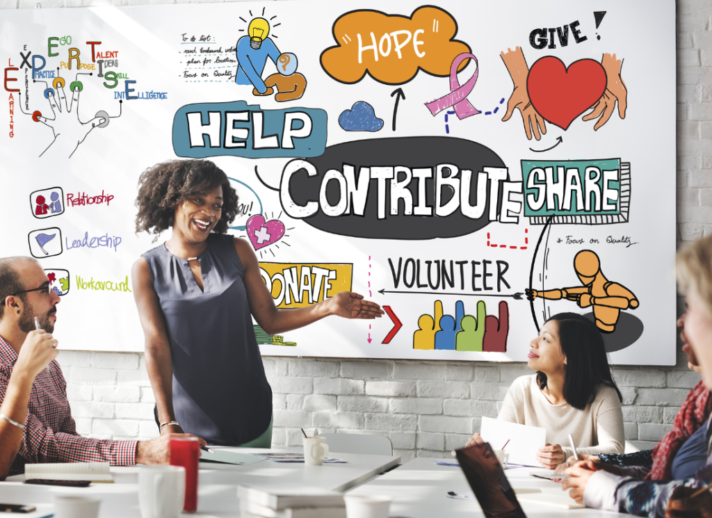 A woman is standing in front of a white board and facing a table of people sitting and listening to her speak. On the white board are graphics and words related to nonprofit services.