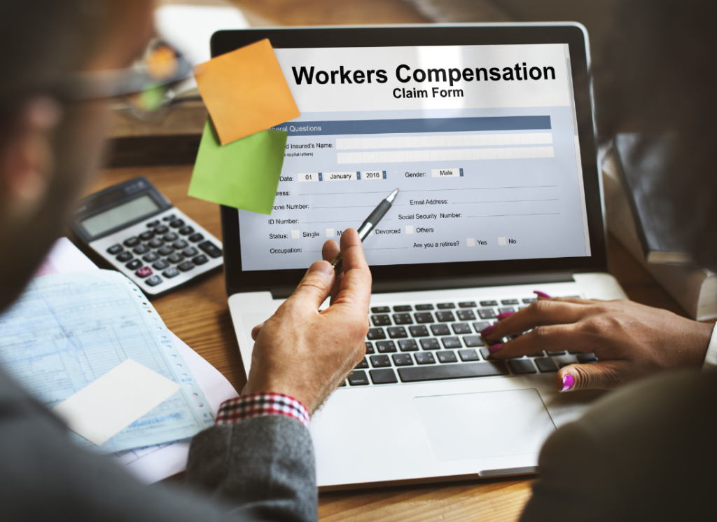 Two people are sitting in front of an open laptop computer. One person is typing while the other is pointing at the screen. The computer is sitting on a desk, and the screen displays a Workers' Compensation form.