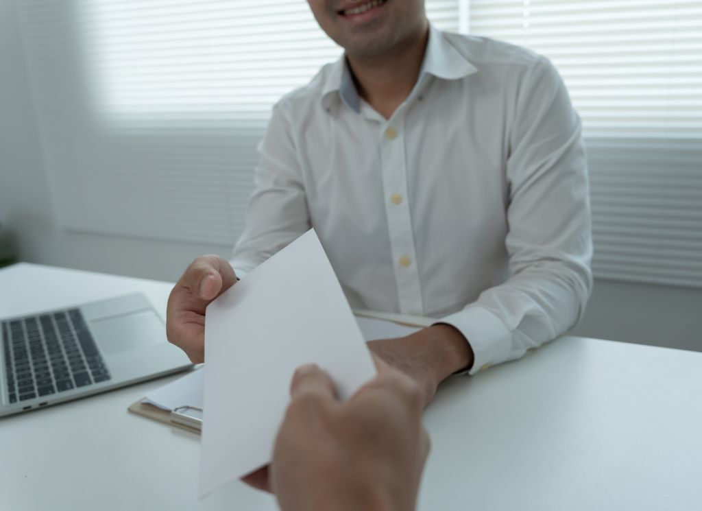 An employee is sitting at a desk in front of a laptop computer and clipboard. The employee is being handed an employee bonus in an envelope.