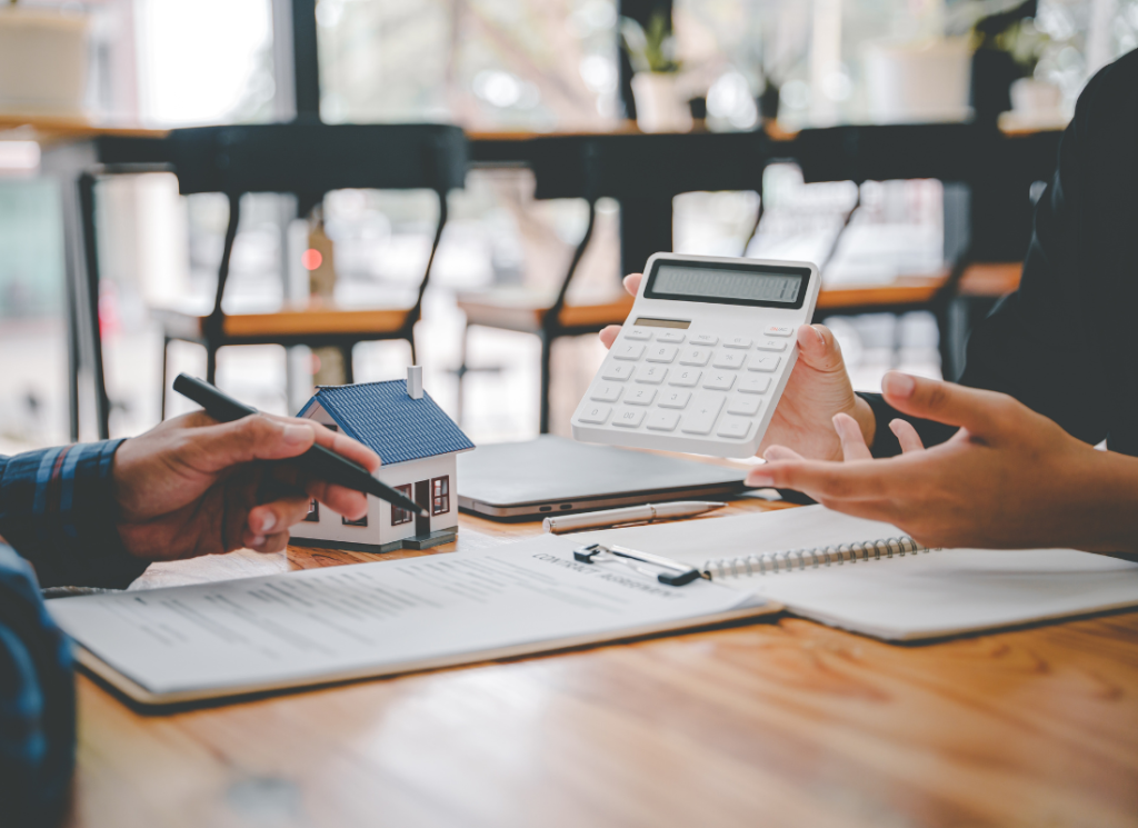 Two real estate professionals are sitting across form each other at an office desk and reviewing documents. One of the real estate professionals is holding up a calculator and showing it to the other person at the desk.