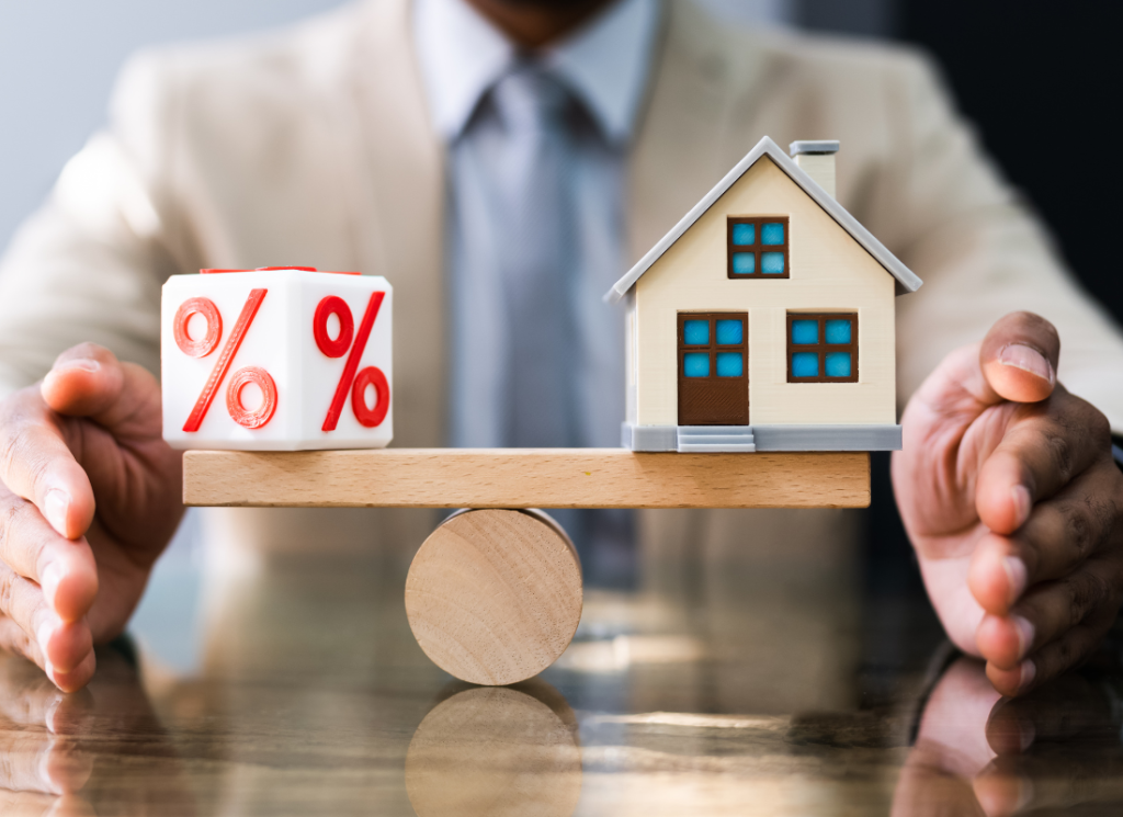 A man is sitting at a desk and balancing a small model house and a block with a percent sign on it.