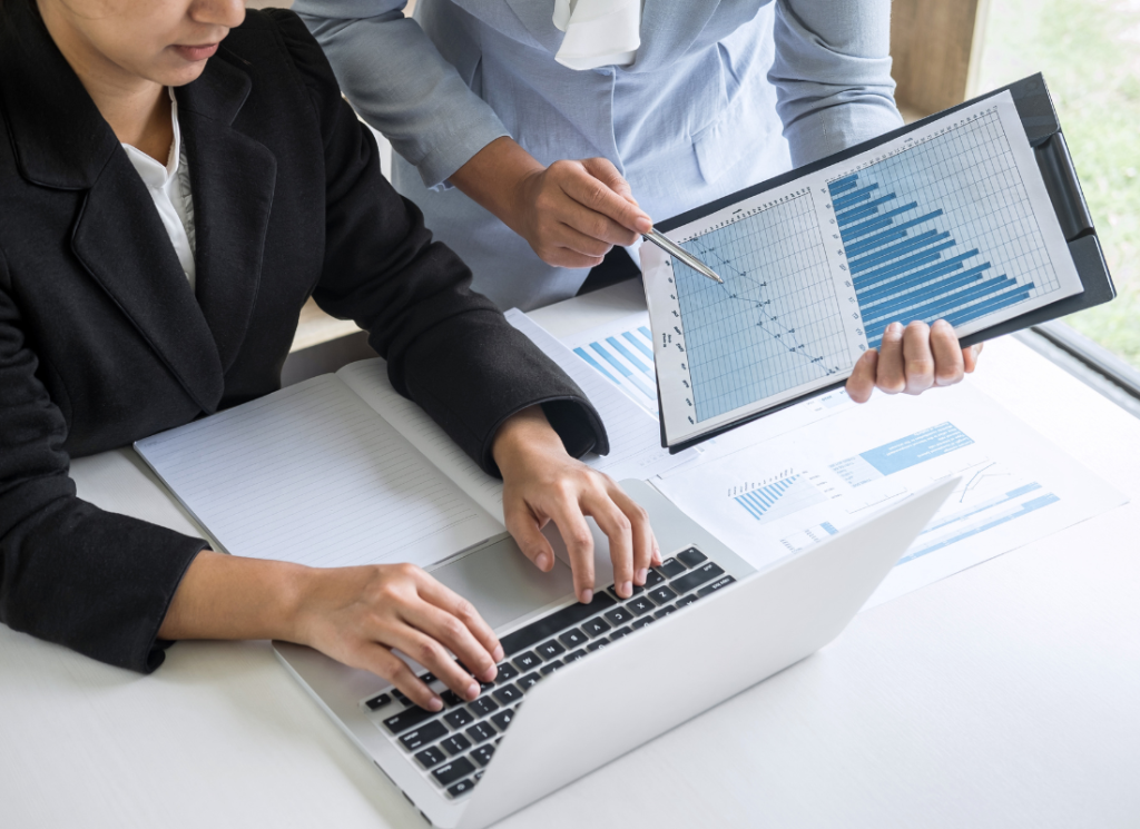 Two people in business roles are at a desk viewing data charts on a clipboard and on a laptop. One business person is pointing to graphs on the clipboard.