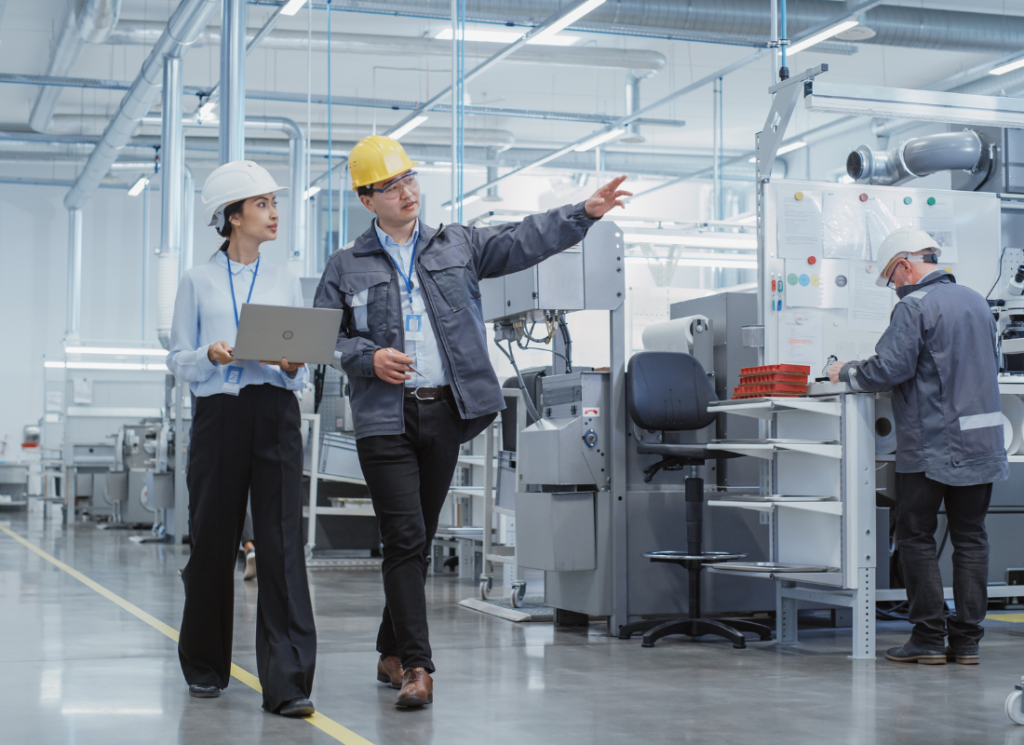 Two employees at a manufacturing company are wearing hard hats and walking while discussing their job. One employee is holding a laptop, the other is pointing to something in the distance.