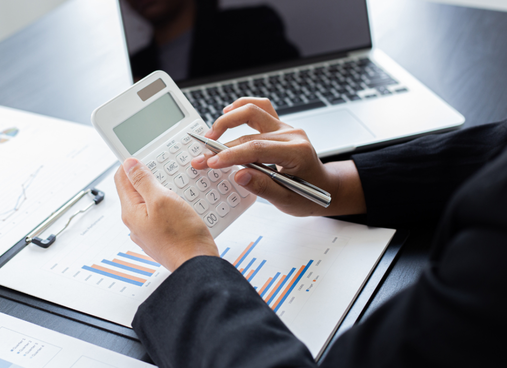 An individual is sitting at a computer desk and holding a calculator. There are printed graphs on the desk in front of the computer..