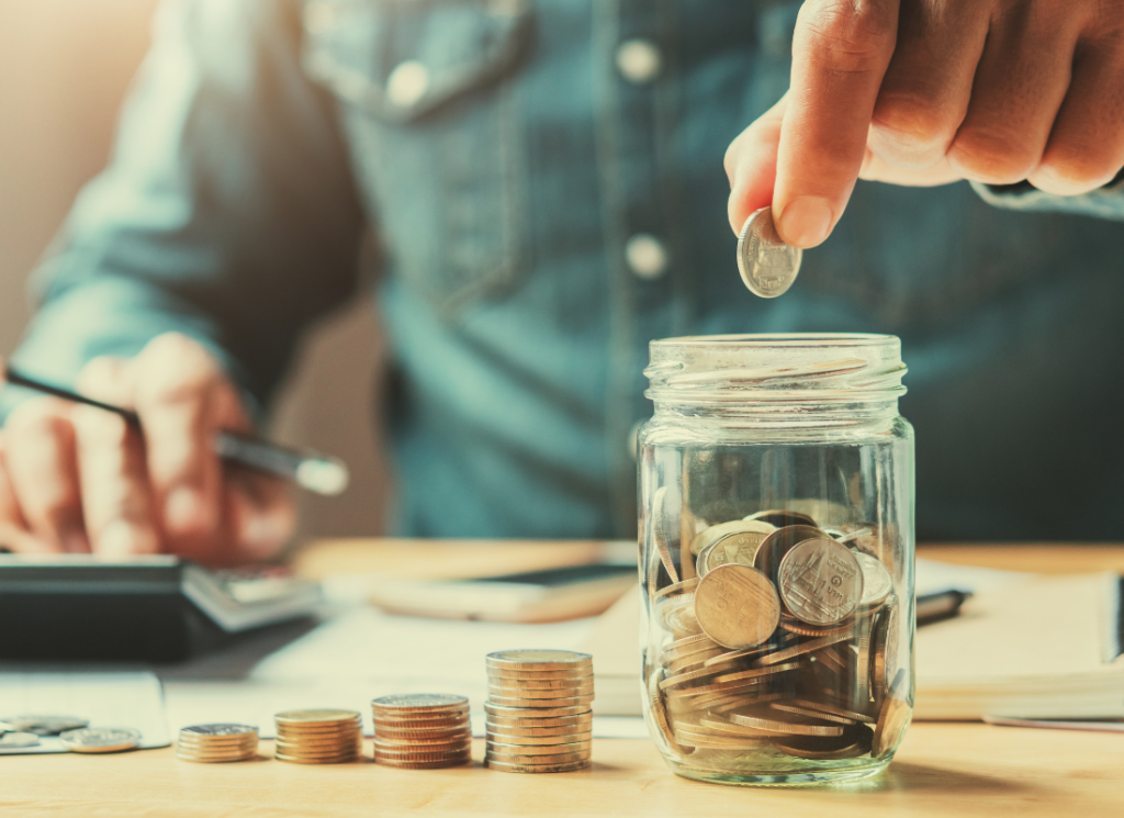 A person is sitting at a desk and placing a coin into a donation jar. The person is holding a pen and typing on calculator at the desk.