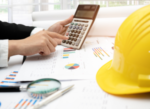 An accountant is working at a desk and typing on a calculator. There are printed documents, a magnifying glass, and a construction hard hat on the desk.