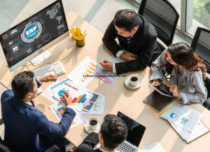 A group of four people are sitting at a table in a conference room and reviewing documents and graphs. There is a computer on the table with 