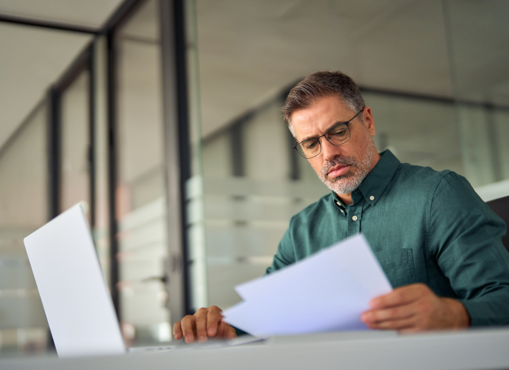 A businessman is sitting at a desk with an open laptop computer. He is reviewing financial statements that he is holding in his hand.