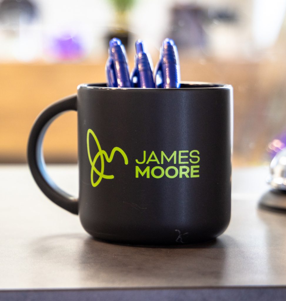 Black coffee mug on a desk with a James Moore logo on it and pens inside the mug.