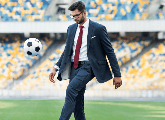 A college athlete wearing a business suit is standing in a stadium and kicking a soccer ball.