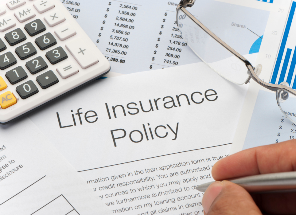 An individual's hand is holding a pen above a life insurance policy. The life insurance policy is on a desk along with a financial document, a calculator and a pair of glasses.
