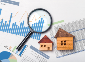 A magnifying glass and two small house models laying on top of multiple graphs, charts, and financial spread sheets.