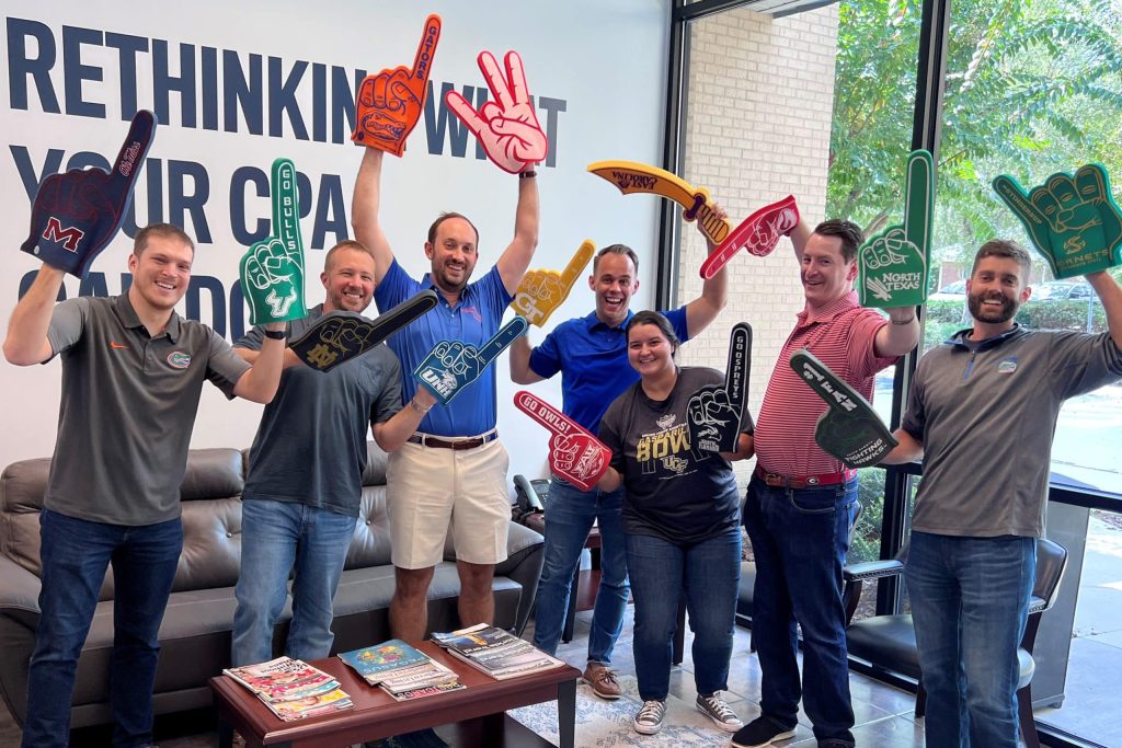 James Moore Gainesville office employees dress in shirts and polo shirts with their favorite college logos on them. Each individual is holding up a foam finger of one of the collegiate athletics clients.