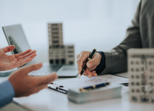 A real estate professional is writing on a clipboard while seated at a desk. There is a second individual seated at the desk as well as two building models, a laptop, and cash.