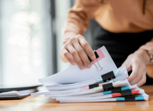 A business person is viewing a stack of documents that are separated by binder clips. The person is in an office setting and the documents are stacked on a desk.