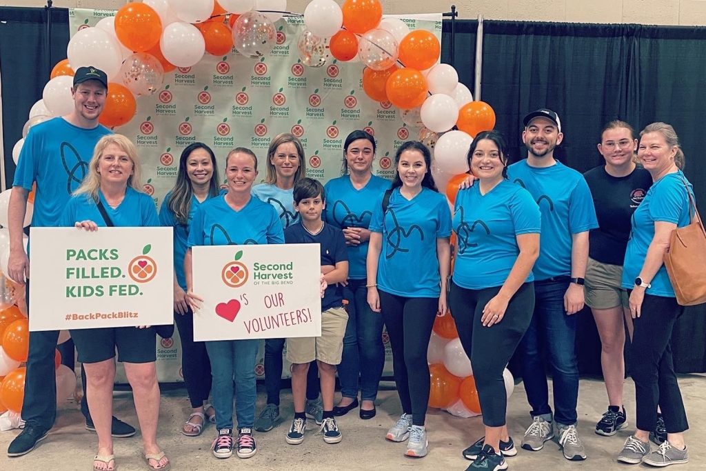 A group photo of James Moore Tallahassee employees at Second Harvest, standing in front of an orange and white balloon background. All the employees are wearing a blue James Moore logo tshirt.