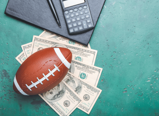 A football lying on top of a small pile of cash. Above the football is a calculator, pen, and padfolio.
