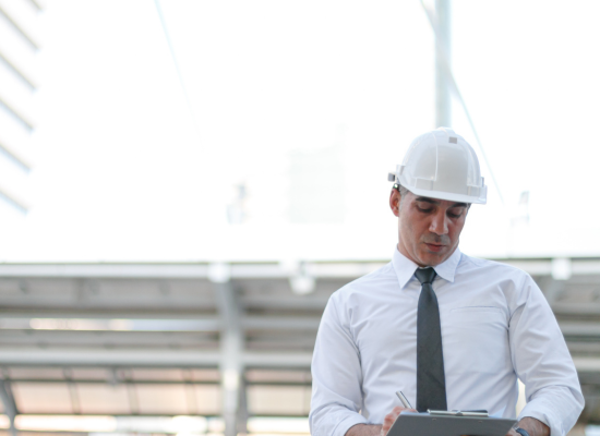 A construction bookkeeper , wearing a hardhat, reviews information on a clip board while at a construction job site.