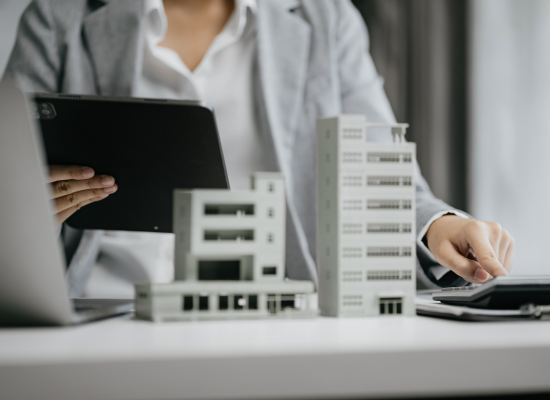 A real estate professional is sitting at a desk reviewing tangible property regulations on a tablet while typing numbers in a calculator. There are two building models on the desk.