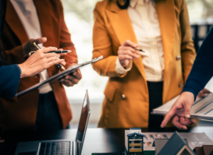 A real estate team engaged in a discussion about balancing capitalization thresholds and debt covenants for their real estate fund. The team is focusing on a building model on a table and a clipboard holding documents.